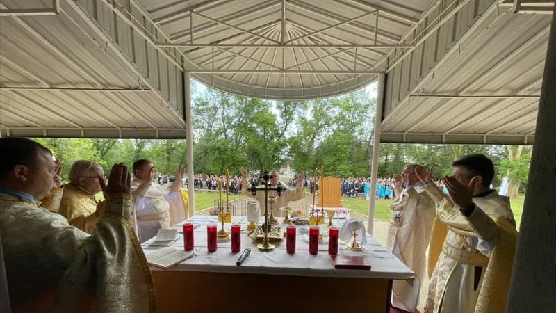 Clergy in vestments raise hands in prayer during an outdoor Divine Liturgy with a congregation gathered.