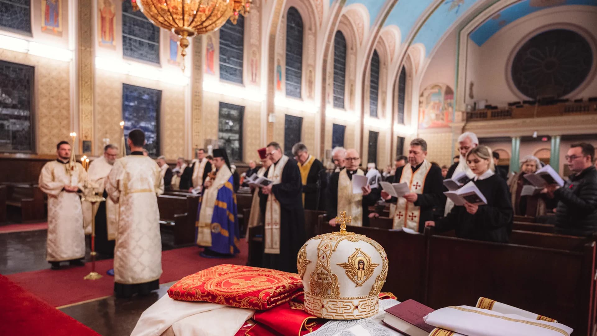 Congregation attends Divine Liturgy in a church with a decorated iconostasis and frescoes.