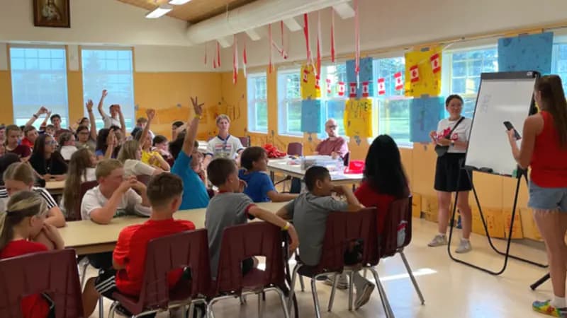 Children and adults raise hands in a classroom with Canadian flags and an icon of Jesus.